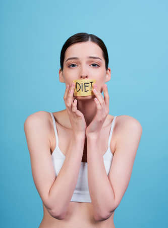 Cute young Caucasian girl in a pink top, holds fork with salad in her hand, looks at her, holds her hand near her mouth, poses in the studio on a blue background. The concept of diet and weight loss.の写真素材