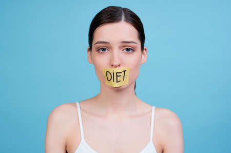 A hungry sad young girl in a white top, holds in her hand a fork with a slice of cucumber and a bowl with green fresh salad, her mouth is sealed with a ribbon with the word food, she is losing weight.の写真素材