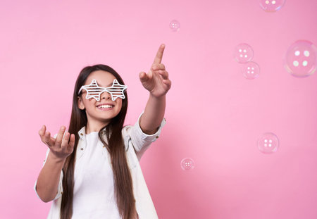Young cute Caucasian brunette schoolgirl with glasses, smiling, holds a book in her right hand in...