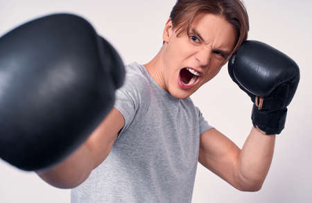 A young aggressive boxer, holding his hands in black boxing gloves near his face, emotion aggression. Caucasian young man boxing, shouting, sports, victory, adrenaline on a white background.の写真素材