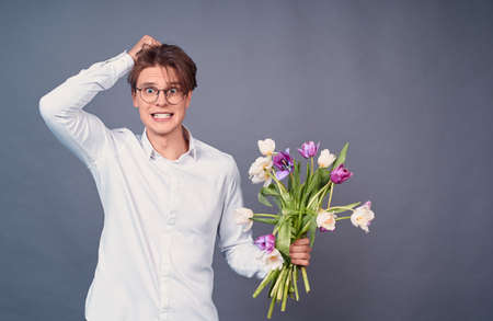 Sad scared guy with a spoiled hairstyle, in a white shirt, holds broken white and pink tulips, looks straight, regrets about a failed date, stands on a gray background.の写真素材