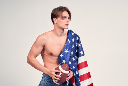 A sexy muscular young American guy, a macho man with a naked torso, an American flag on his shoulders, holding an American football ball in his hand, looking to the side. Copy space.の写真素材