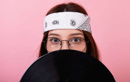 Emotional young long-haired girl hipster in a bandana, glasses,a vest on a pink background. She covers her face with a retro vinyl disc, a plate for listening to hit songs. Musical concept.Copy space.の写真素材