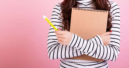 A slender long-haired young brunette girl in a striped blouse with a pen and a notebook presses to herself to gain new knowledge on a pink background. Learning concept. Copy space.の写真素材