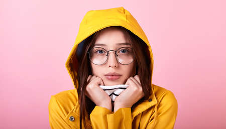 A cute Caucasian girl in a yellow rain jacket and a hood and a striped blouse with glasses on a pink background. A lady hugs herself and shakes from the cold. She is sad on a rainy day. Copy space.の写真素材