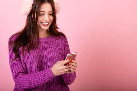 Sweet girl in a knitted sweater and fluffy pink headphones holding a phone. Looks and smiles at the phone. Photo on a pink background. Winter mood, winter clothes. New Year's bustle.Copy space.の写真素材