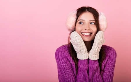 Funny young lady in a knitted sweater and mittens and fluffy winter pink headphones. The girl smiles looks to the side and presses her hands to her face close-up. Photo on pink background. Copy space.の写真素材
