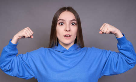 Surprised young Caucasian girl keeps herself fit and healthy, raises her hands and shows muscles, dressed in a blue sweatshirt, stands on a gray background.の写真素材