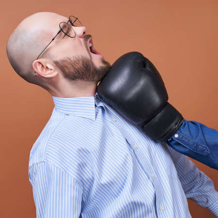 Close-up, a young handsome man in round glasses and a light business shirt, stands in profile, receives a blow to the jaw from the side with a hand in a black boxer glove.の写真素材