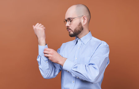 A handsome young man, stands in profile, he has a gorgeous beard and round glasses, buttons a button...