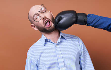 A funny young businessman with a beard, in a blue shirt and round glasses, shocked by the sudden blow of a boxing glove from the side.の写真素材