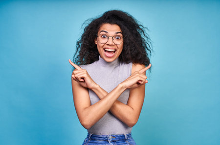 Cute cheerful dark-skinned woman with curly hair, posing against a blue background, shows direction...