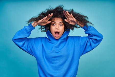 A surprised, shocked, discouraged young African American woman, wearing a blue hoodie, holds her black curly hair in her hands. The concept of unruly hair, afro hairstyle.の写真素材
