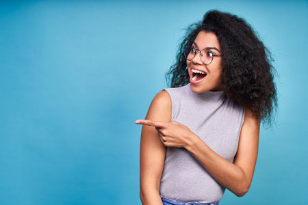 Horizontal shot of a beautifull young joyful African American female point with front finger off, points to copy space to left on blank wall, isolated on blue background. Copy of space.の写真素材