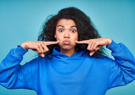 Funny dark-skinned curly woman points her finger to her cheeks, makes grimaces, wears a casual blue...