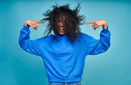 Funny dark skinned curly woman points her fingers at her fluffy black curly hairstyle, wears a casual blue sweatshirt, posing against a blue background, copy space.の写真素材