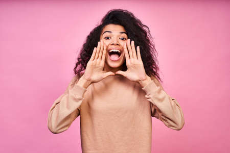 A cute stylish young dark-skinned woman, with beautiful dark skin, black curly hair, holds her palms near her mouth and shouts loudly, wears a brown blouse and stands against a pink background.の写真素材
