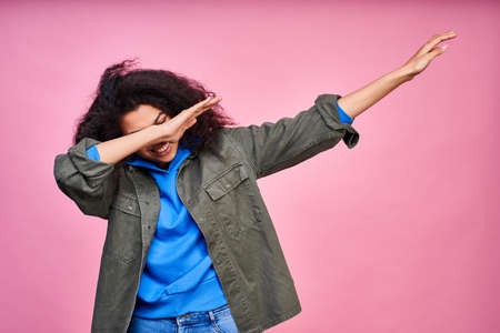 Funny happy young dark-skinned girl, with lush curly black hair, wears everyday clothes, makes hands dance gestures, having fun on a pink background.の写真素材