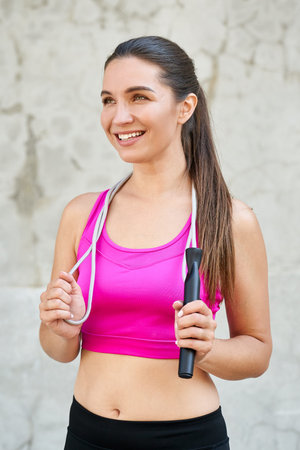 Vertical shot of a smiling, happy, affable athletic girl, wearing a pink top, holding a rope around...
