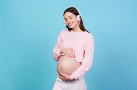 Pregnant cute young woman listens to music on headphones on a blue background and keeps her hands on her stomach. The concept of pregnancy, motherhood, preparation and expectation.の写真素材