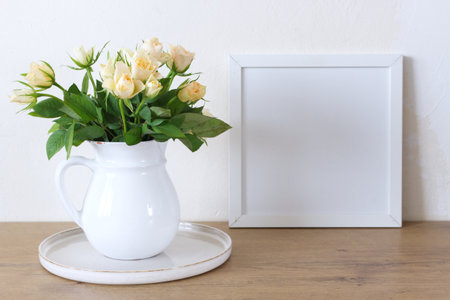 Square wooden frame mockup on a vintage bench, table. Modern white ceramic vase with white roses. White wall background. Scandinavian interior. Roses concept.の写真素材