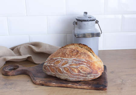 Breakfast still life. Fresh baked surdough bread on wooden chopping board. Kitchen interior. White tiled background. Food concept. Milk can.の写真素材