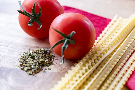 Dry spaghetti, fresh tomatoes and Italian herbs, closeup, high angle. Ingredients of Italian pasta meal, red napkin, wooden background.の写真素材