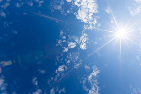 A bright sun with white rays departing from it. In the blue sky there are clouds illuminated by sunlight.の写真素材