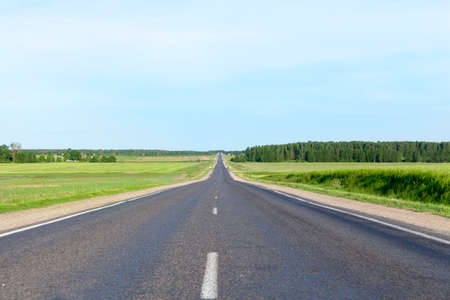 A small rural asphalt road. Landscape with blue sky, grass and trees. On the roadway cars are movingの写真素材