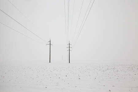poles with electric lines in the winter season. cloudy weather with white sky and white snow on the ground. landscapeの写真素材