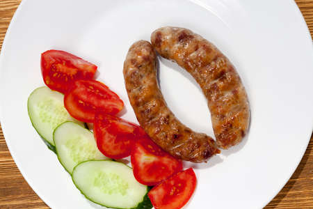 Two meat pork sausages, fried on a barbecue, cucumbers and tomatoes, lying on a white plate after cooking. photo close-up, top view.の写真素材