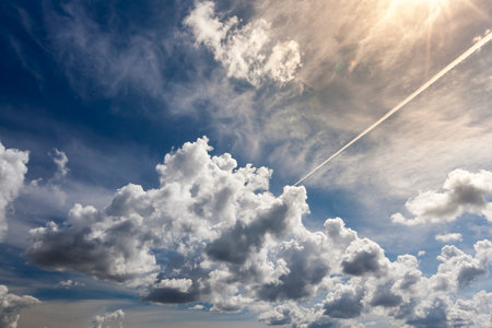 huge cumulus clouds and a white long trail from an airplane in a blue bright sky, several rays of...