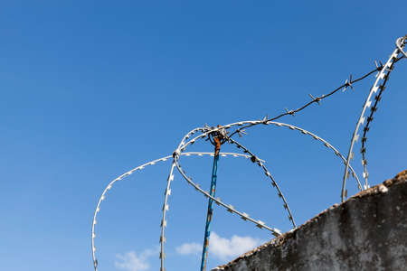 old dirty and high concrete fence on top of which is wound a new metal wire to protect the territory, against a background of blue sky close-upの写真素材