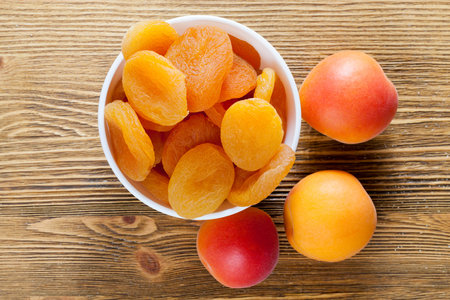 fresh dried apricots in a white bowl and fresh fruit lying together on a wooden table, closeupの写真素材