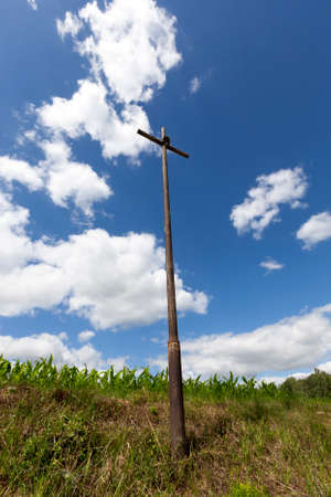 a tall metallic religious old cross set on a hill in a field, against a blue cloudy sky and green grassの写真素材