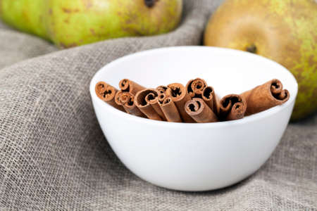 whole cinnamon sticks, lie in a white deep bowl next to green pears, on a linen tableclothの写真素材