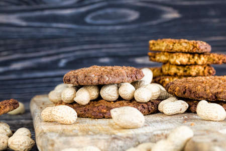 A stack of oatmeal cookies with crushed roasted peanuts and lying on a wooden table next to whole nuts of inshell peanutsの写真素材