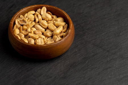 peeled peanuts in a wooden round bowl, crunchy peanuts without shells on a black slate board,...