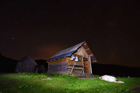 Beautiful landscape with a house in the mountains against the sky with stars and a dog in the grass in the yardの写真素材