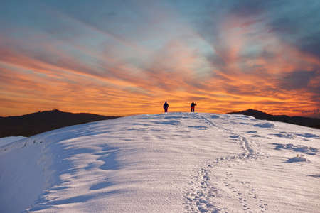 Two tourists walk on a mountain slope to the summit against the backdrop of mystical clouds at sunsetの写真素材