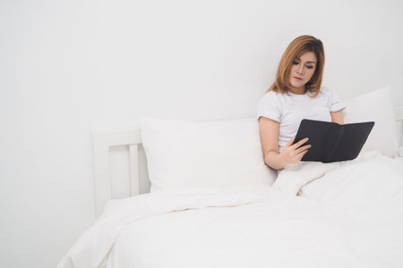 Asian woman reading book on white bed, left copy space area.