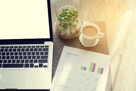 office table with blank screen on laptop, cup of coffee, garden plant in glass pot and statistic report paper at living room, concept of business lifestyle. selective focus on top view.の写真素材