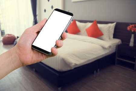human hand hold smartphone, tablet, cell phone with blurry modern bedroom. concept of  finding hotel.の写真素材
