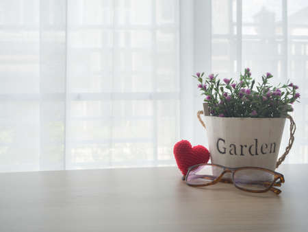 wood office table with pink rose flower on pot, red heart shape sign and modern eyeglasses on beautiful white drape window texture background at living room.の写真素材