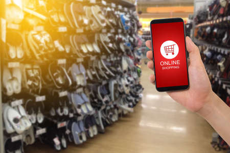 human hand hold smartphone, tablet, cell phone with shop cart online shopping on blurry footwear shop, Online shopping concept.の写真素材