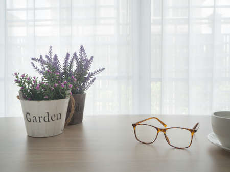 office table with beautiful variety flower on pot and modern eyeglasses, white ceramic coffee cup on blurry beautiful white drape window texture background.の写真素材