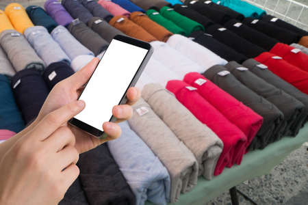 human hand hold and touch smartphone, tablet, cell phone with blurry shelf of colorful T-shirt on market.の写真素材