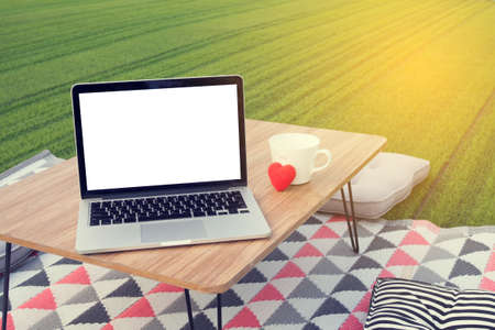 picnic table with blank screen on laptop and white coffee cup, red heart shape at green lawn garden. concept of workplace in holiday. view from front notebook screen.の写真素材
