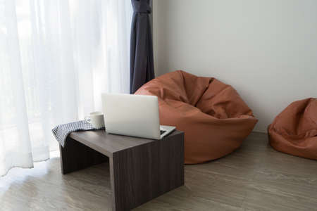 Office table with laptop, white coffee cup and orange beab bag on white curtain windows texture background in living room. view from front table.の写真素材