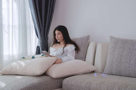 sad asian woman looking out side and using smartphone, cellphone on sofa at living room. concept of young woman broken heart.の写真素材
